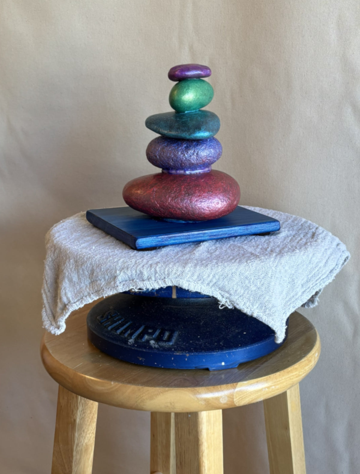 Stacked stone sculpture by Robert Schott with beautiful jewel colored stones sitting on a platform which itself is sitting on a stool. 